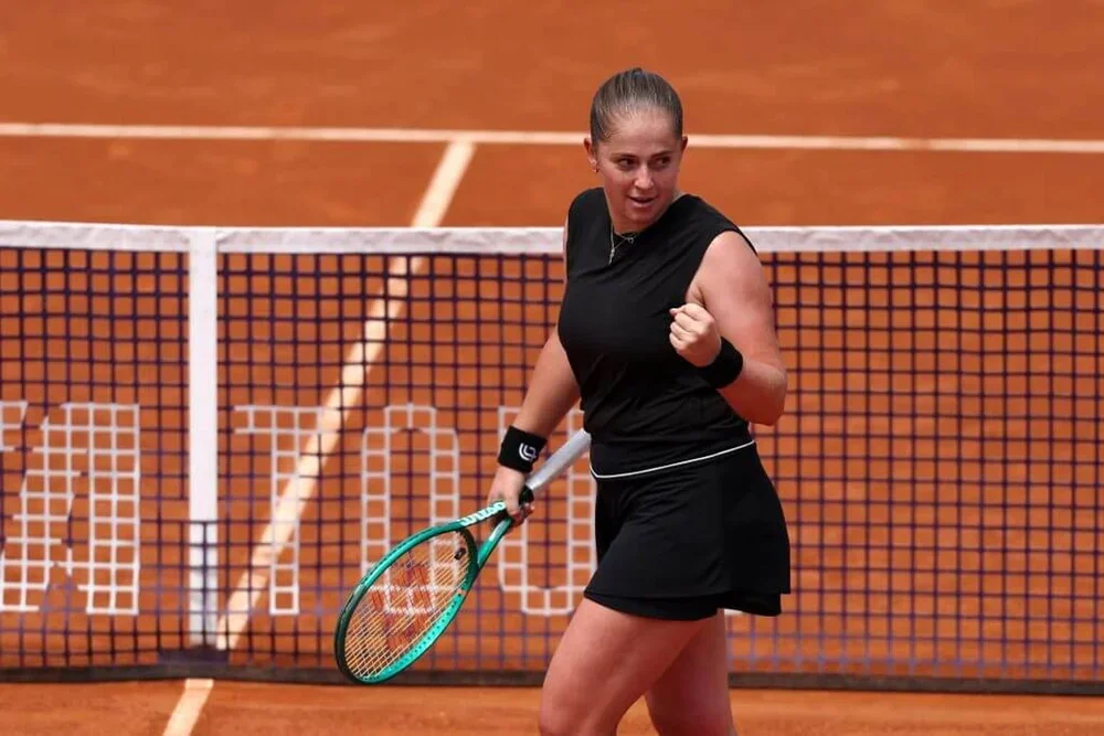 Jelena Ostapenko, standing on a clay tennis court, turns to her left and pumps her left fist while holding a green tennis racket in her right hand.